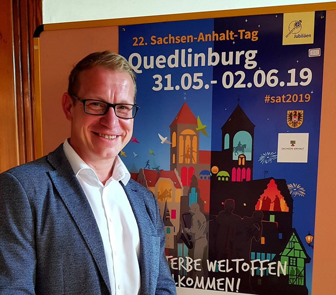 Das Bild zeigt Norman Groß vor dem Siegerplakat zum Sachsen-Anhalt-Tag 2019 in Quedlinburg.